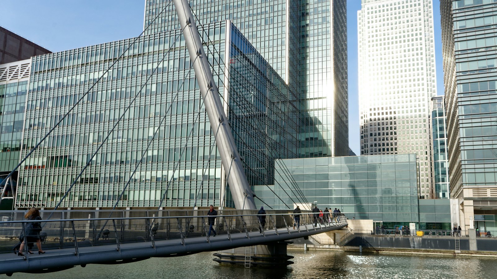 Una pasarela con un mástil blanco inclinado y cables de acero sobre el agua en Canary Wharf, Londres, en un día soleado, con personas cruzándola y torres de oficinas de cristal al fondo