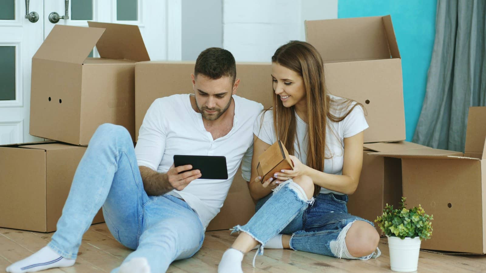 Dos personas sentadas en el suelo entre cajas de cartón de mudanza, una con una tableta y la otra con una pequeña caja de madera
