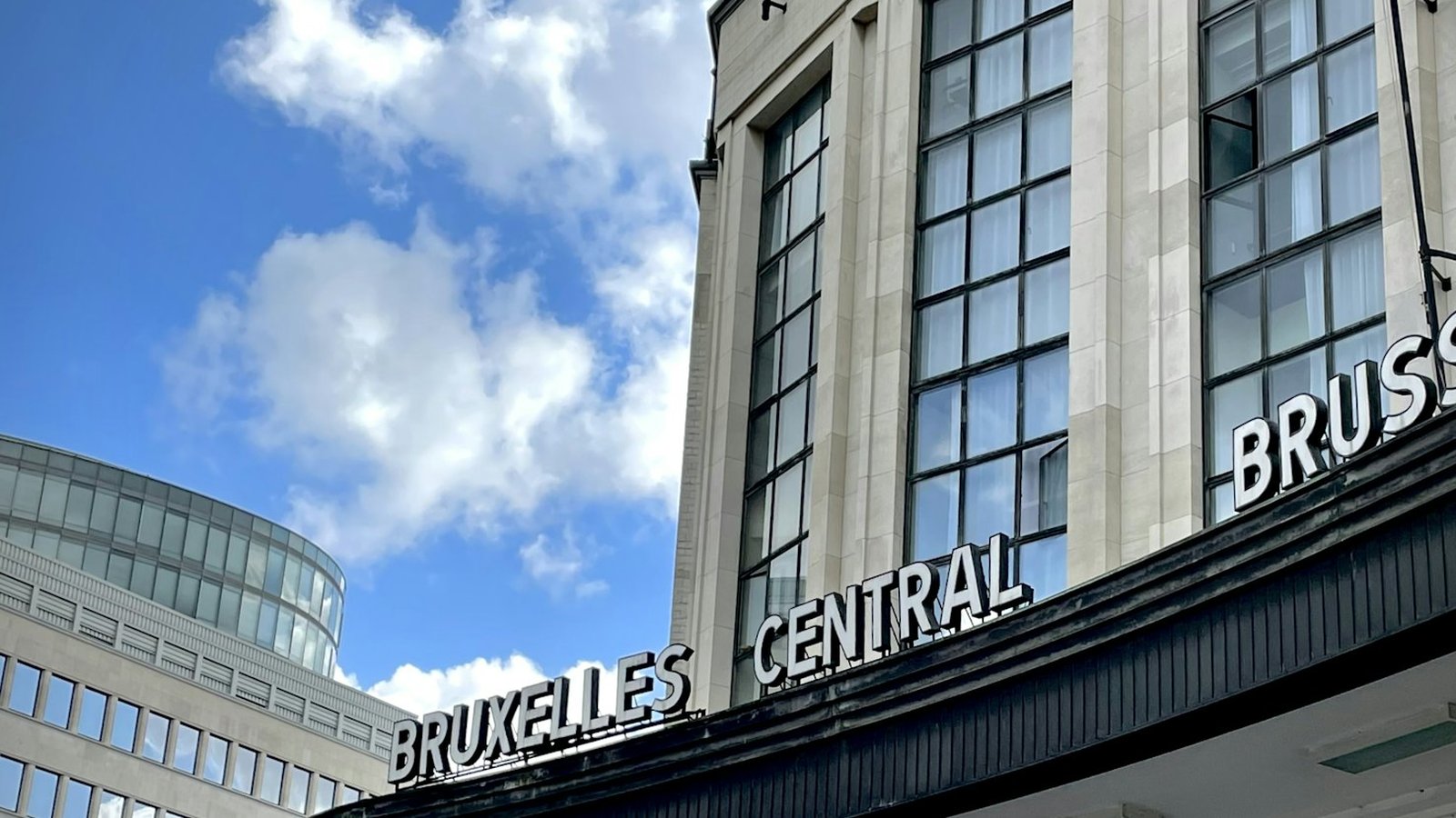 The facade of Brussels Central station under a blue sky with white clouds, with the station name in French, Bruxelles Central, in large white letters and the start of the Dutch name beside it