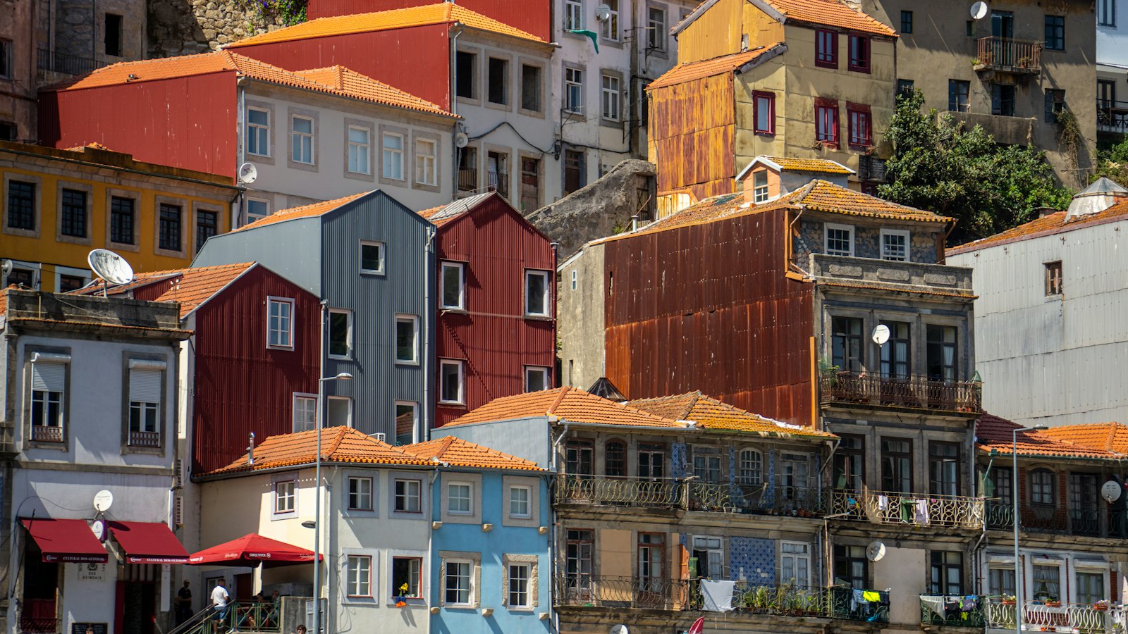 Case addossate su un ripido pendio di Porto in una giornata di sole, con tetti in terracotta, facciate in lamiera ondulata rossa, blu, gialla e grigia, balconi in ferro con il bucato steso e tende di un caffè al livello della strada