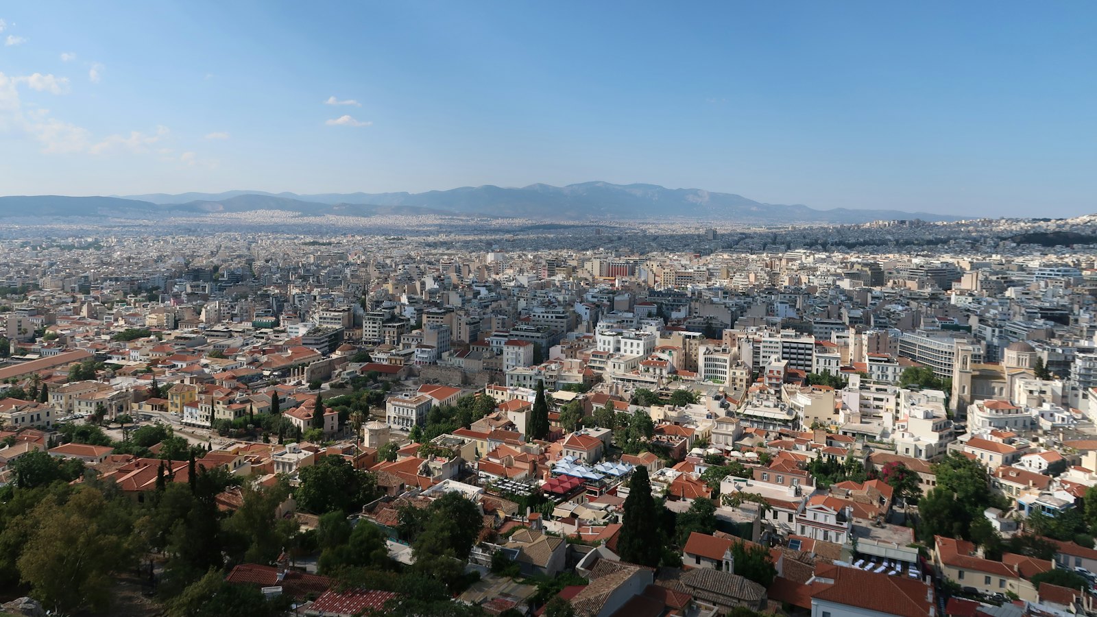 Atene vista dall'alto in una giornata limpida, con tetti di tegole rosse e alberi in primo piano, isolati fitti di palazzi bianchi fino all'orizzonte e una catena di montagne sullo sfondo