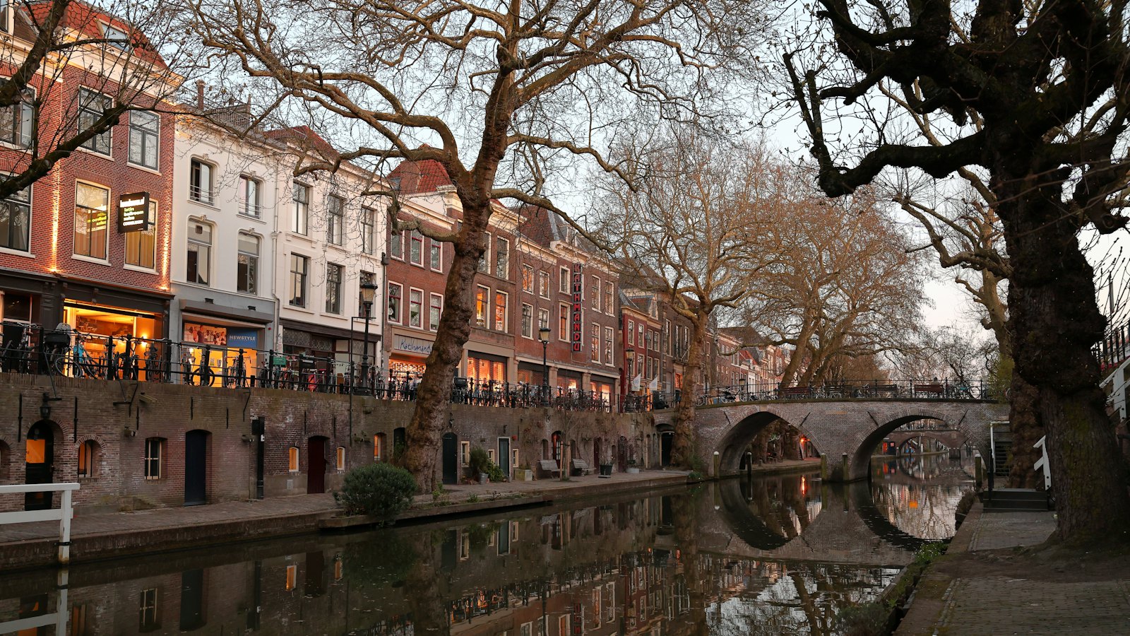 Il canale Oudegracht a Utrecht al tramonto, con le cantine in mattoni delle banchine a livello dell'acqua, vetrine illuminate e platani spogli lungo la banchina, e un ponte ad arco in mattoni riflesso nell'acqua ferma