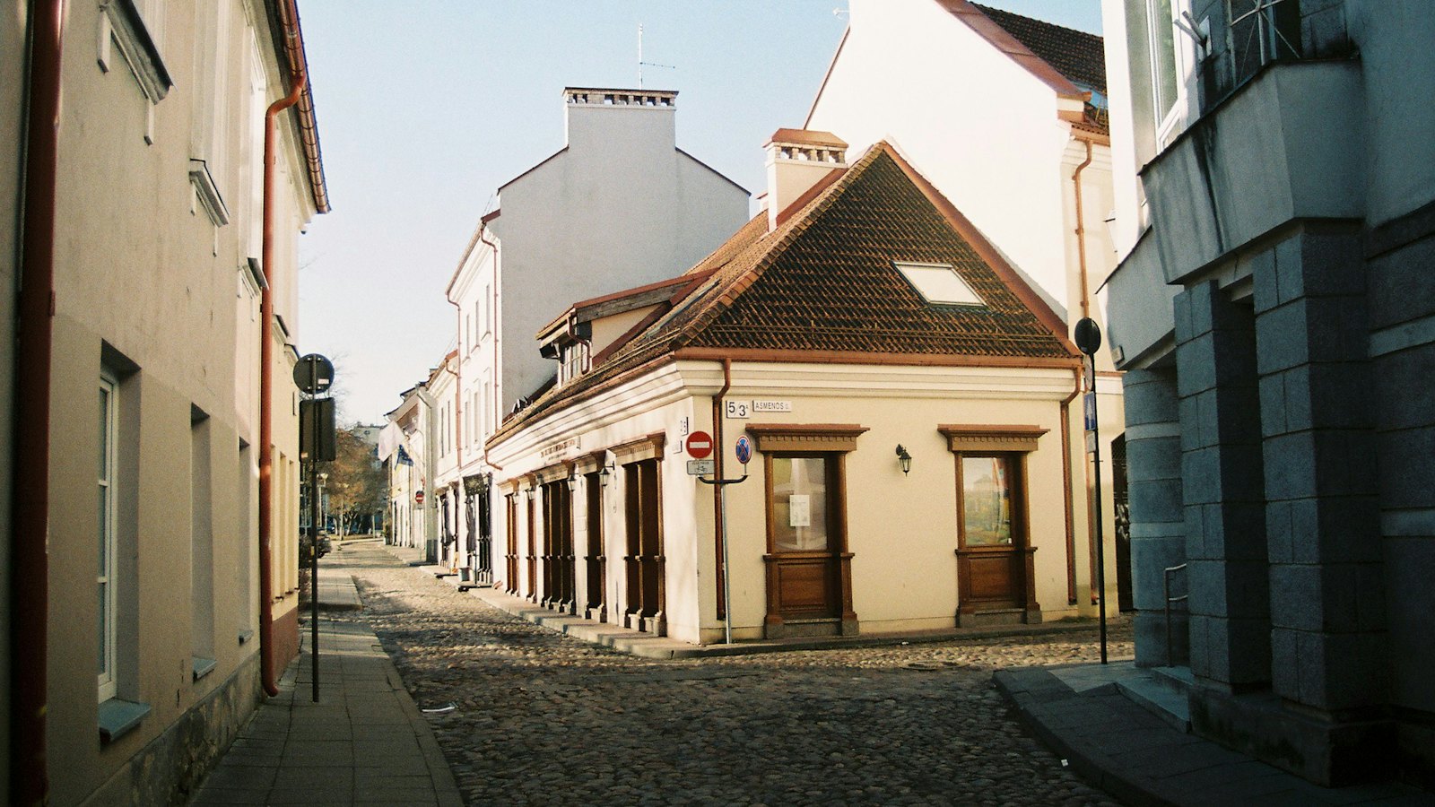 Una tranquilla strada acciottolata nel centro storico di Vilnius, con edifici bassi dai toni chiari e una casa d'angolo con tetto di tegole e finestre in legno