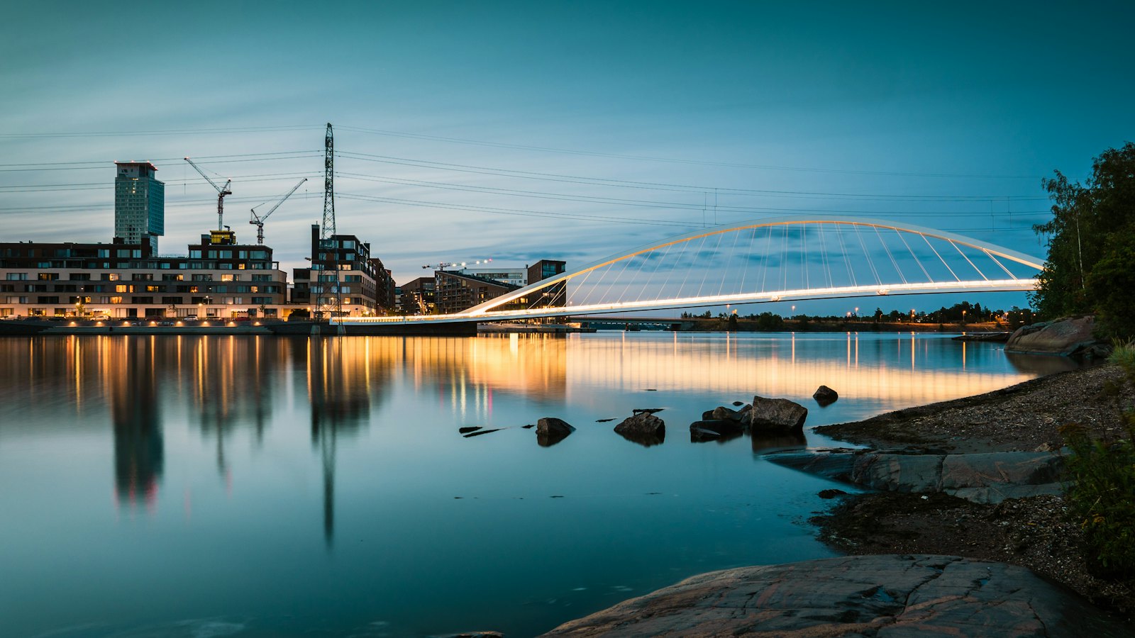 El arco blanco del puente Isoisänsilta en Kalasatama, Helsinki, iluminado al anochecer, con bloques de viviendas, grúas y agua en calma que refleja las luces